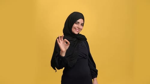 Fröhliche muslimische Frau in schwarzer Kleidung, die das OK-Zeichen mit den Händen zeigt und lächelt, Erfolg und gute Laune