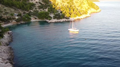 Aerial fly-by of drone over cove in the mediterranean sea with sailboat and Island in the setting su