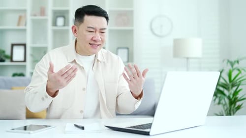 Angry excited asian businessman arguing over video call using laptop while sitting in home office.