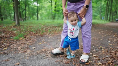 Mom’s holding her baby’s hands helping him to walk. Little child learning to walk outdoors.