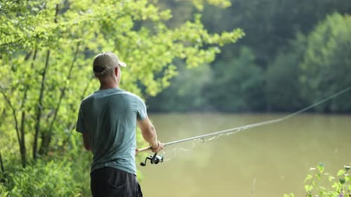 Male angler gets his fishing rod out in murky lake surrounded by trees on sunny day