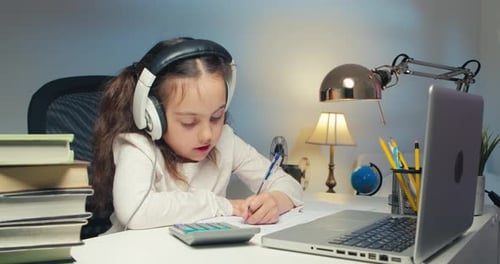 Little Girl in Headphones is Studying Online at a Laptop at Home
