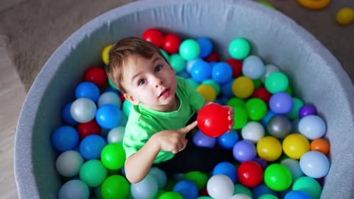 Cute toddler boy sitting in the pool with ball. Kid holds a red ball and pokes it with finger.