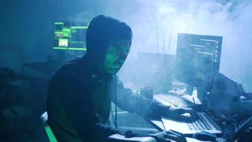 Un mystérieux hacker travaille dans une pièce faiblement éclairée entourée de plusieurs écrans d'ordinateur