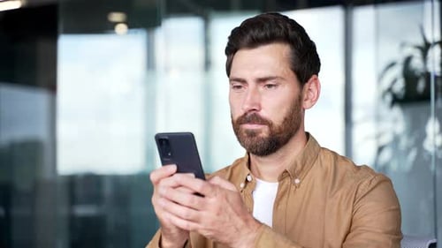 A smiling businessman is using browsing mobile phone at the workplace in a modern business office.