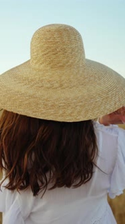 Femme au chapeau de paille traversant un champ rural ensoleillé, brise calme et paysage ouvert et chaud