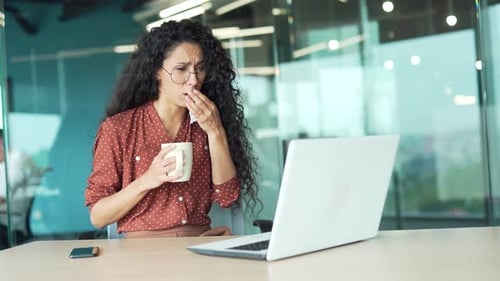 Sick young businesswoman suffers from a cold sitting at a workplace in a business office. Female
