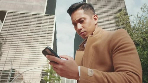 Close up, young businessman is sitting on bench in the street and typing on mobile phone