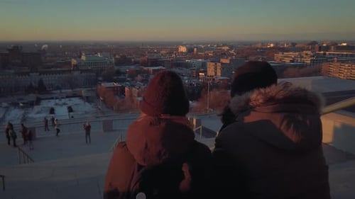 Friends bundled in heavy coats and wearing masks sit outside on steps of building enjoying sunset du