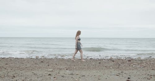 Young adult caucasian woman walking on the beach near sea on cloudy day, full length female outdoors