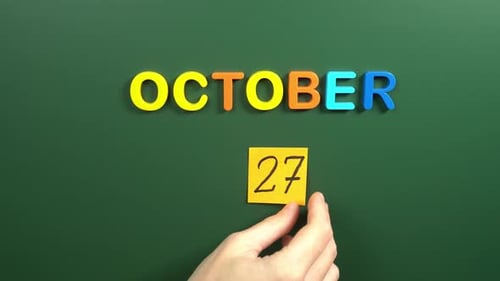 Hand sticking a sticker 27 October calendar day on school board. 27 date of October. Twenty seventh