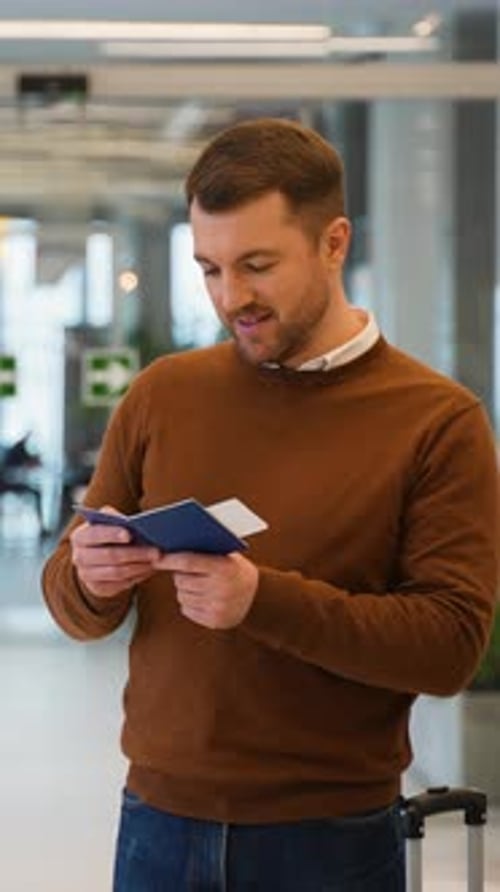 Young Man Holding Passport and Tickets Looks at the Information Board of Departures and Arrivals at