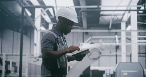 Factory Worker in Hard Hat Uses Handheld Device While Monitoring Robotic Equipment