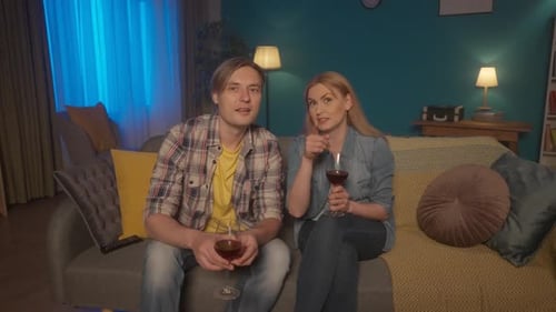 A Couple is Sitting on a Sofa with Glasses of Wine Watching TV A Man and a Woman are Watching a