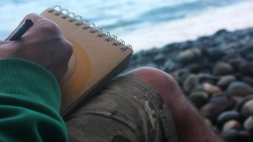 Man doing color pencil drawing in brown paper sketchbook by the sea