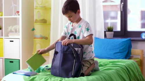 Happy young schoolboy packing schoolbag with school supplies at home before school