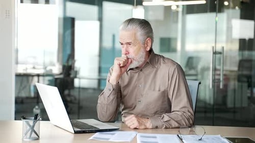 Senior gray haired bearded businessman coughing sitting at workplace at desk in business office.