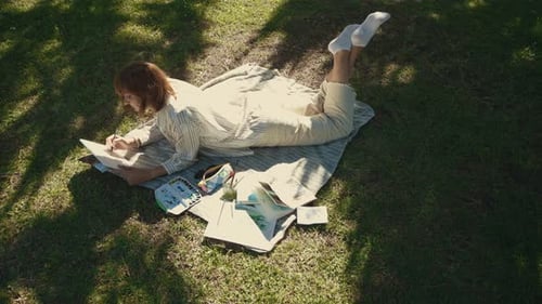 An Adult Inspired Woman Lying on the Blanket in the Green Park and Drawing in Her Sketchbook
