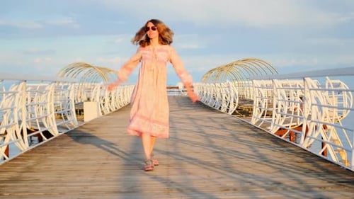 A woman in long dress and sunglasses dance walking on wooden pier on seashore.