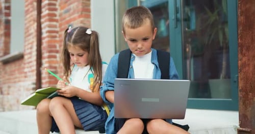 School small students friends outdoors using laptop computer . Kids sitting on stairs and play