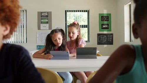 Diverse girls tapping tablet, leaning in and pointing, collaborating in class on math task at table