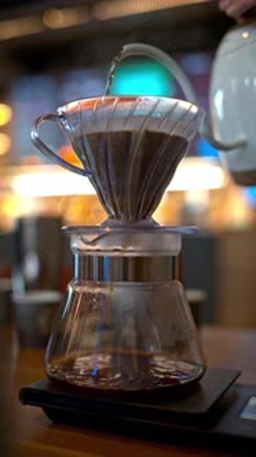 Close up of a man brewing pour-over drip coffee in a slow, circular motion on a wooden table. Vertic