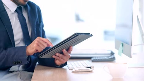 Closeup of businessman holding tablet, sitting and working at desktop computer in modern startup