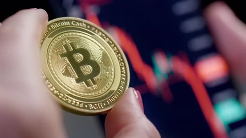 Main féminine aux ongles rouges tenant un bitcoin brillant devant un graphique flou montrant une marque d'ours