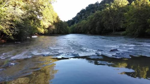 Drone flies low over a river with a reflection of the trees on either side of the river.