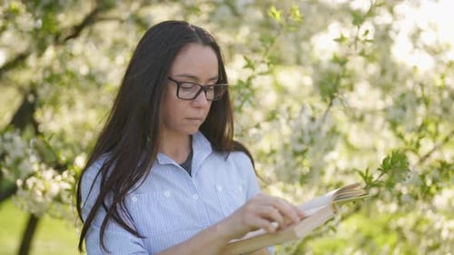 A Stylish Middleaged Woman with Glasses is Standing in the Park By a Flowering Bush and Reading a