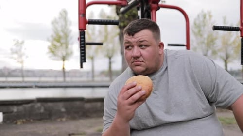 A Very Fat Man Sits on an Outdoor Exercise Machine and Holds a Bun