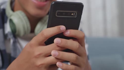 Close Up Of Asian Teen Boy's Hands Playing Smartphone While Sitting On Sofa In The Living Room