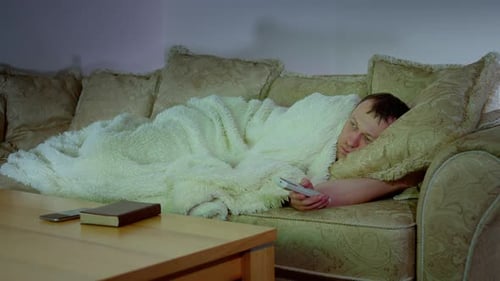 A Man Lying on the Sofa at Night in Front of the TV with the Remote Control
