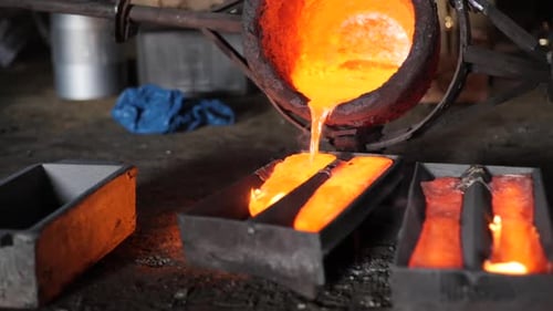 Slow motion foundry bucket pouring orange metal into moulds with flames in a rustic factory setting.