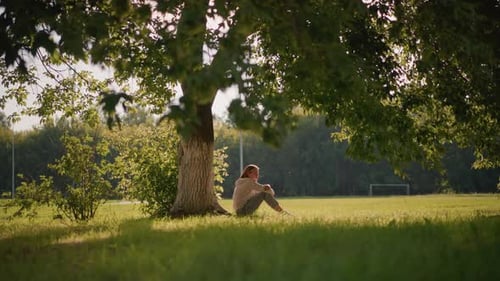 Thoughtful Woman Seated Under Tree in Sunlit Park with Greenery