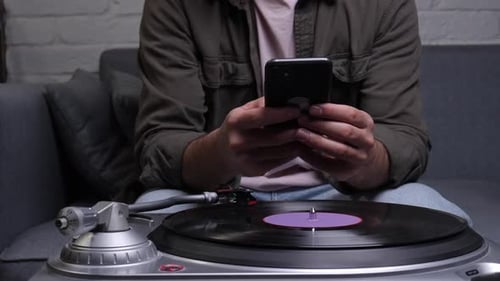 Man Sitting on a Couch Relaxing at Home Using a Smartphone While a Vinyl Record Spins on a Turntable