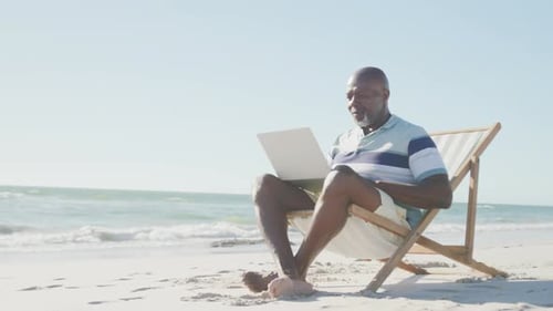 Senior african american man on deck chair, using laptop at beach, copy space, in slow motion