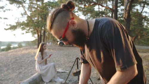 A Young Hipster Man in Shorts Cuts Meat for a Picnic with Friends on the River Bank on a Summer Day