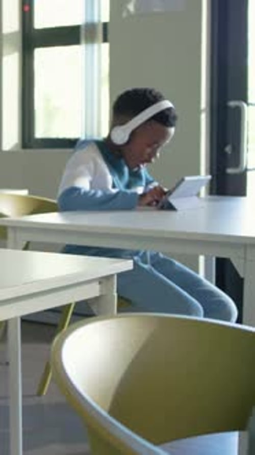 Vertical video: Tapping male student wearing headphones swiping tablet at school desk studying