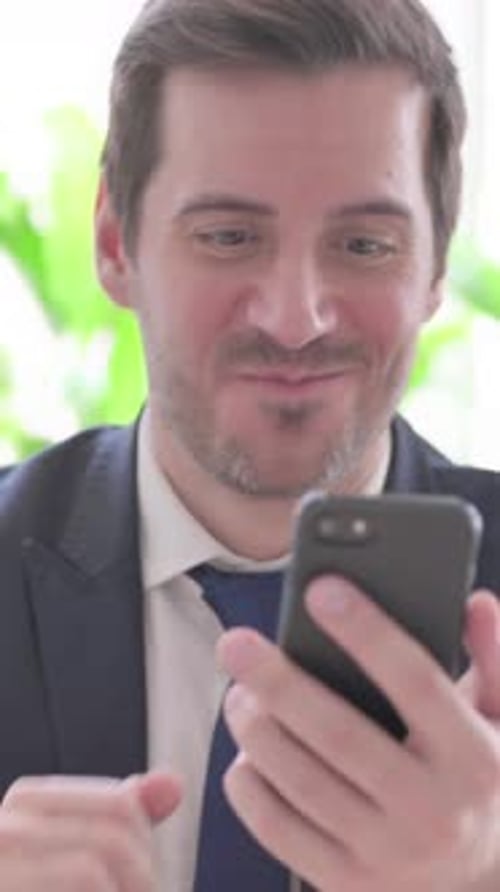 Young Businessman Celebrating Online Success on Smartphone in Office, vertical video