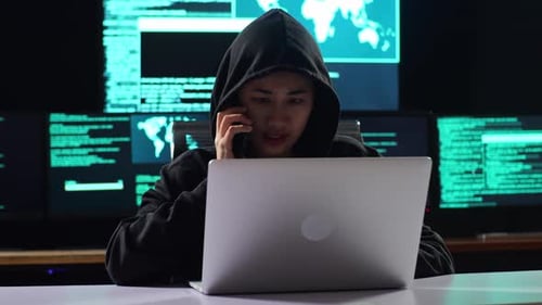 Hacker asiática com capuz falando no smartphone enquanto trabalha em um laptop com vários monitores