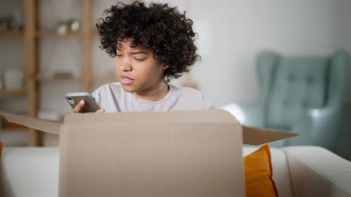 Woman Using Phone While Unpacking Box at Home