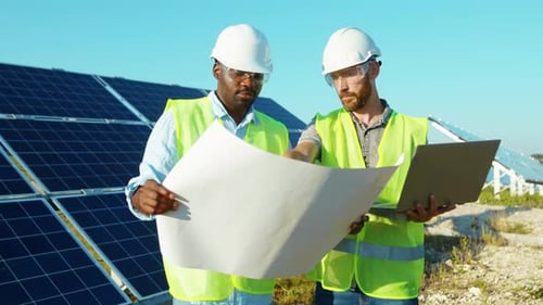 Men Engineers Use Laptop and Project Plan on the Solar Farm in Special Uniform Standing Discuss the