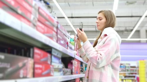 Young Woman Choosing Curling Iron for Hair in Home Appliances Store Goods for Beauty and Hairstyle