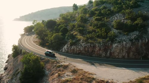 Car with Paddleboard on the Roof Drives Along a Narrow Coastal Road Under a Clear Blue Sky