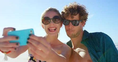 4k video footage of a young couple standing together on the beach and taking selfies on a cellphone