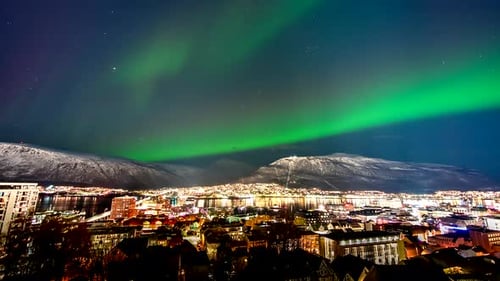 Green aurora borealis dance in night sky over illuminated Tromso city, timelapse