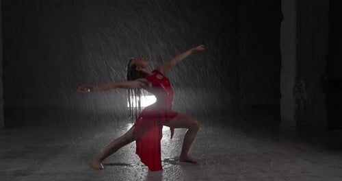 Tempting Young Woman Dancer in Red Dress Performing in the Rain in Dark Studio