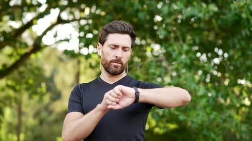 Sports man stands in urban city park and looking smart watch. Handsome bearded athlete using fitness