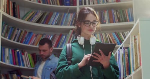 Caucasian Young Female Student in Glasses Standing in Library Using Tablet Device and Tapping on